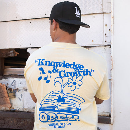 Person wearing a yellow t-shirt with blue graphics and text, standing in front of a white wall.