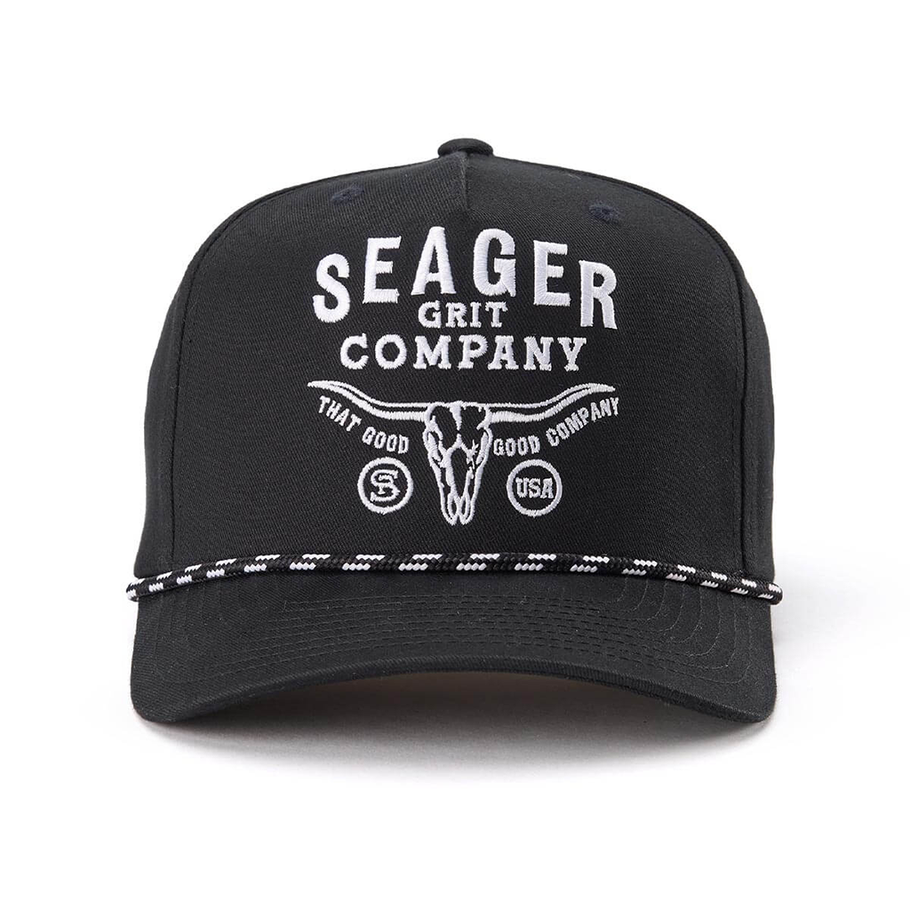 A black baseball cap featuring white embroidered text that reads 'SEAGER GRIT COMPANY' with a longhorn skull graphic and 'THAT GOOD COMPANY' below. A black-and-white patterned rope detail runs across the brim.