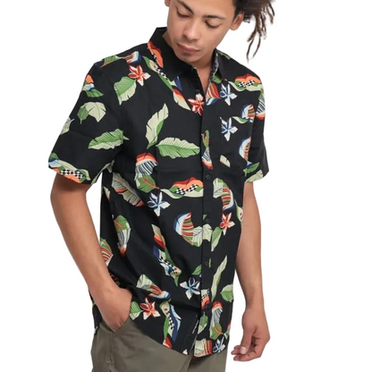 A man wearing a black short-sleeve button-up shirt with a colorful tropical leaf and bird pattern, paired with olive green pants, set against a plain white background.