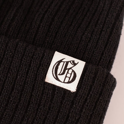 Black knit beanie with a white logo patch on a beige background