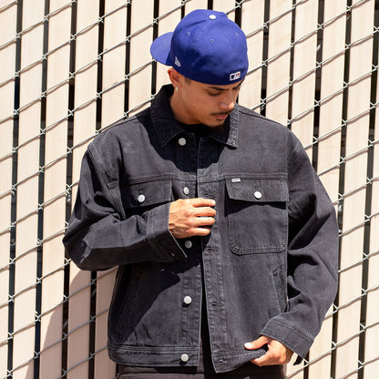 Person wearing a dark denim jacket and blue cap against a wire fence background