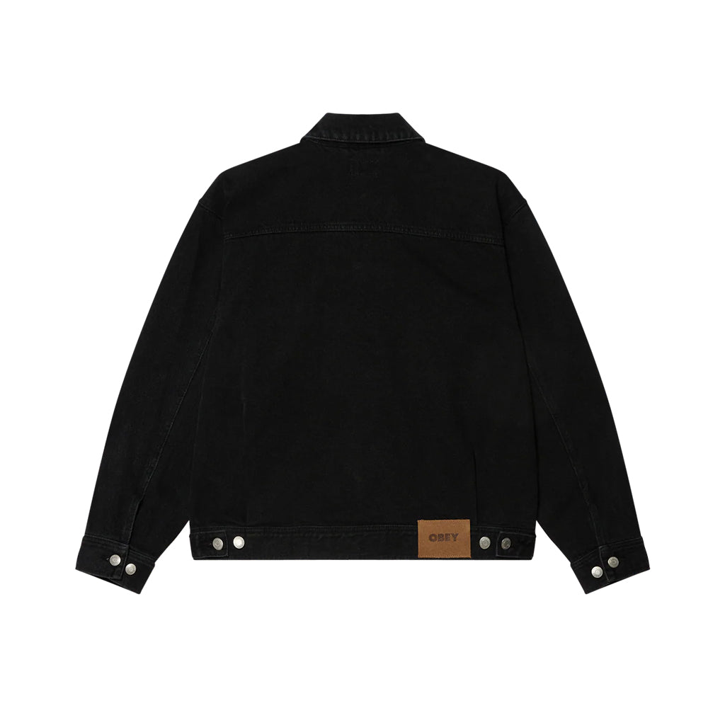 Back of faded black denim trucker jacket with brown obey patch on bottom of jacket. Laying flat on a white background