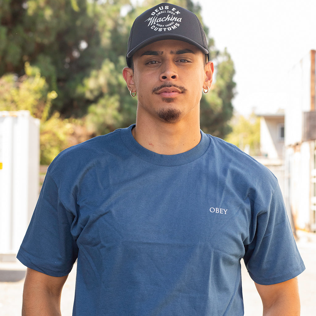 Man wearing a blue t-shirt with 'Obey' logo and a black cap outdoors