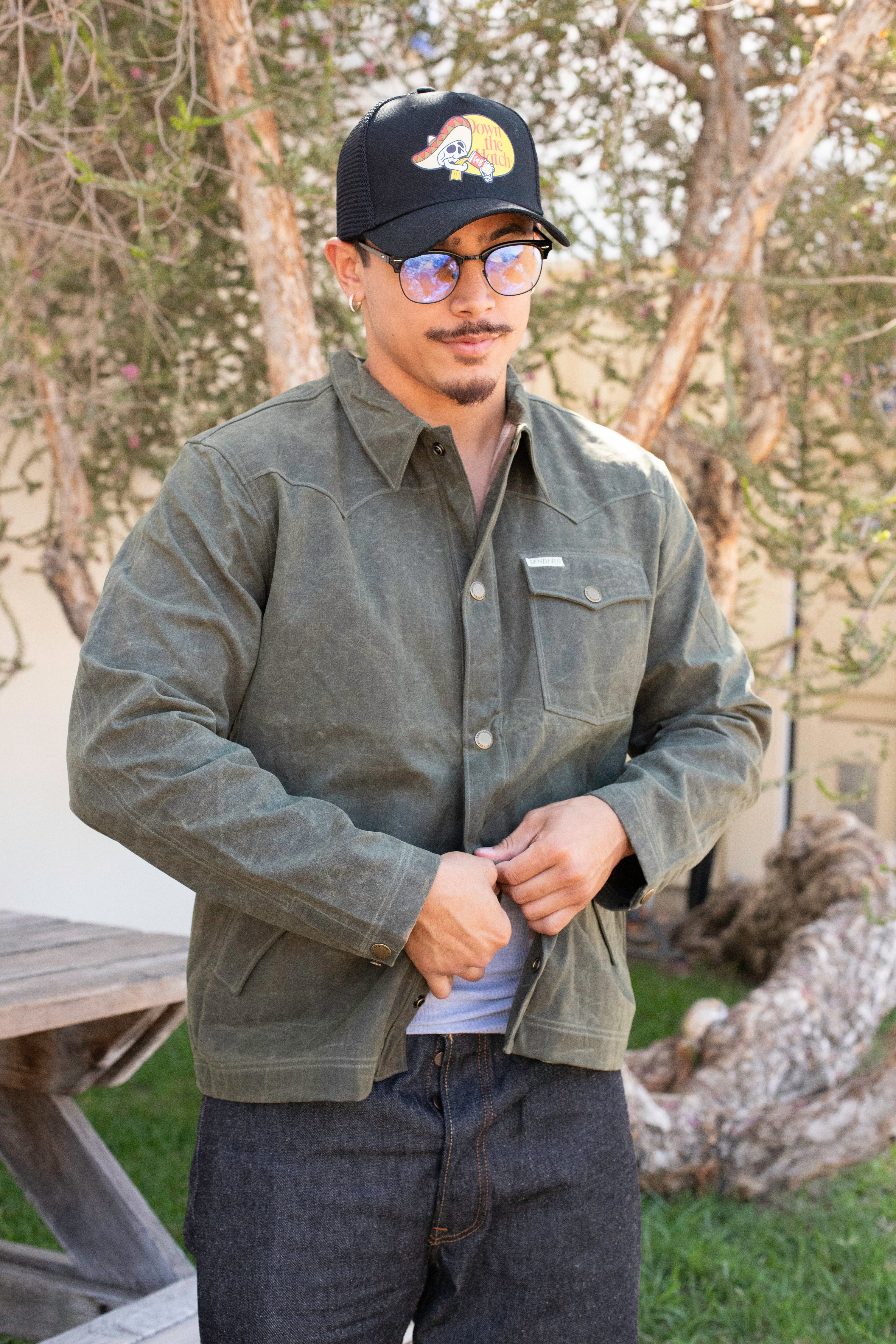 Man wearing a green jacket and black cap outdoors with trees in the background