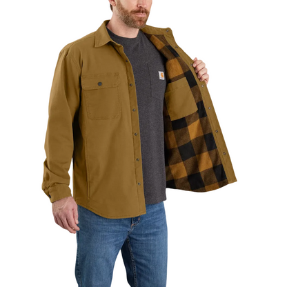 A man wearing a Carhartt work shirt in a camel brown color, shown with his left hand opening the collar to reveal a black and brown buffalo plaid lining. He is also wearing a dark gray t-shirt and blue jeans, set against a plain white background