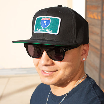Person wearing a black cap with a 'Santa Ana' patch and sunglasses, standing against a neutral background.