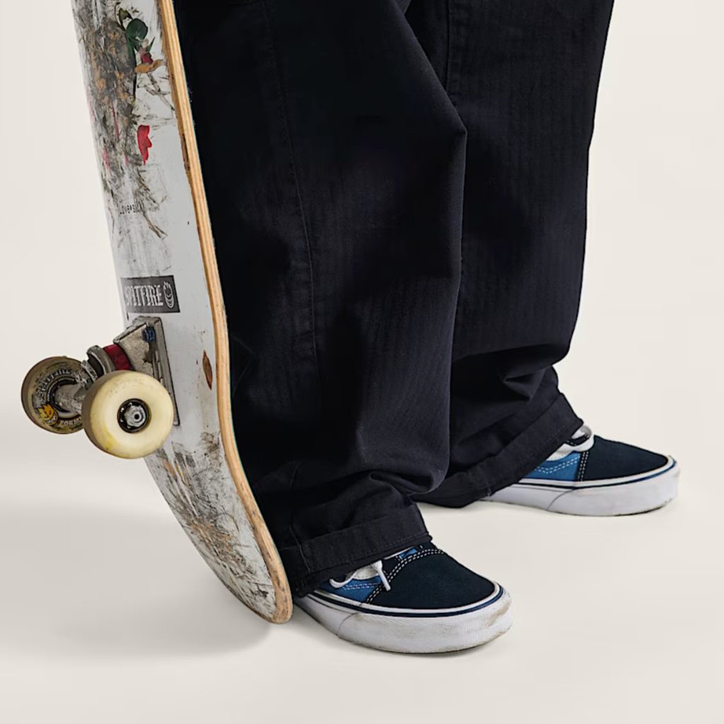 Person wearing black pants and blue sneakers standing next to a skateboard with a colorful design.