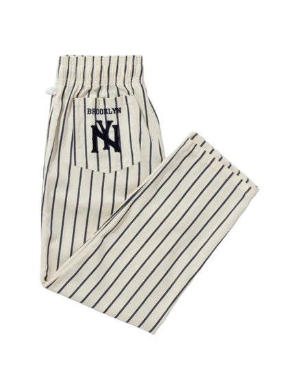 A pair of pinstriped lounge pants featuring a back pocket with a navy blue 'BROOKLYN' text and 'NY' logo patch. The pants are off-white with thin dark vertical stripes.
