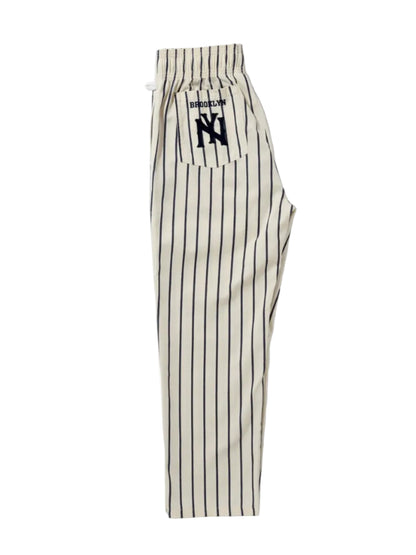 A pair of cream-colored pinstriped pants with black vertical stripes, featuring a drawstring waistband and a back pocket with a black embroidered 'Brooklyn' 'NY' logo. 