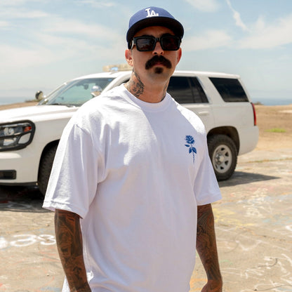 Man wearing a white t-shirt with a blue logo, sunglasses, and a cap in front of a white SUV.