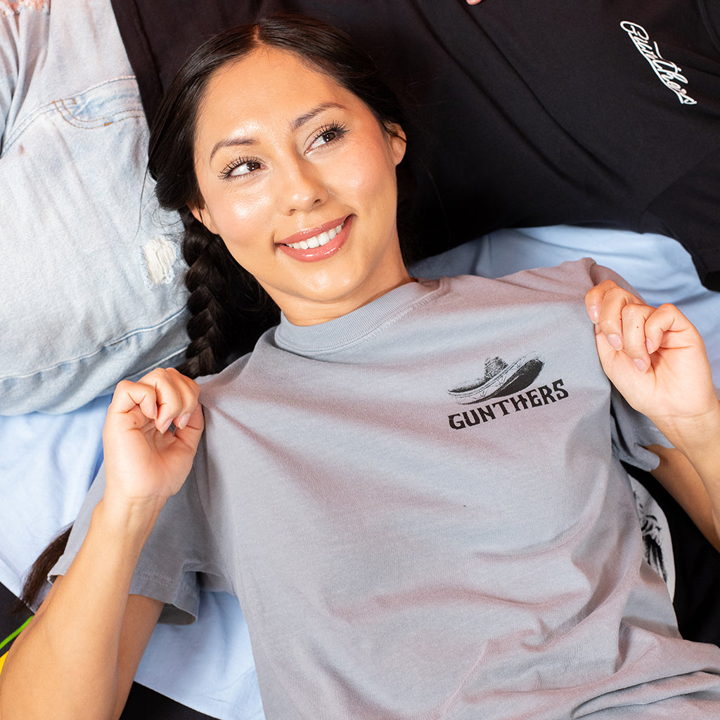 Woman holding up a gray t-shirt with 'Gunthers' logo
