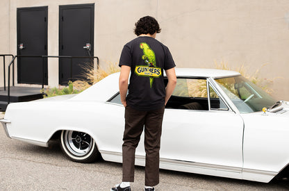 Person wearing a black t-shirt with a green parrot design, standing next to a white classic car.