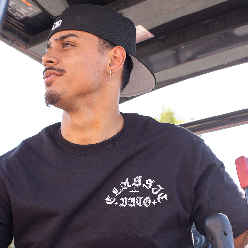 Man wearing a black cap and black t-shirt with 'CLASSIC VATO' text, sitting inside a vehicle.