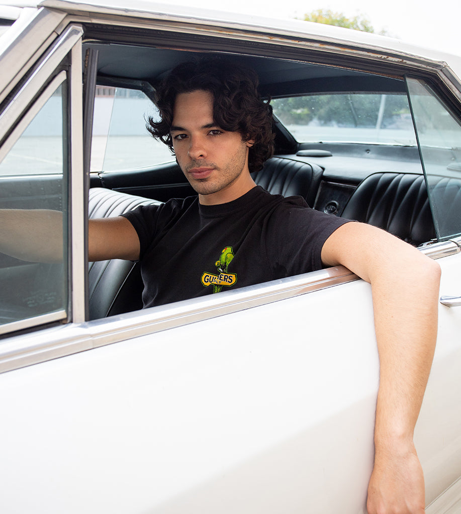 Man sitting in a classic car wearing a black t-shirt with a logo.