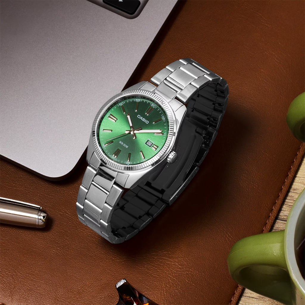 Silver watch with a green face on a brown surface with a laptop and mug.
