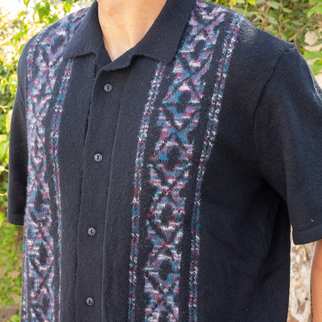 Person wearing a dark shirt with a patterned vest over it, outdoors.