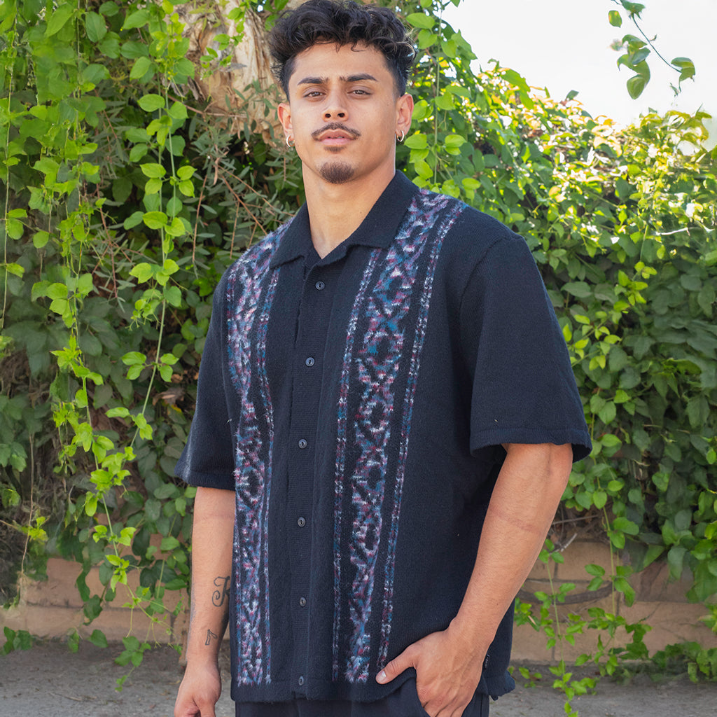 Man wearing a dark shirt with a colorful pattern standing in front of green foliage