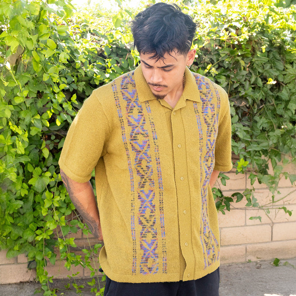 Man wearing a mustard yellow shirt with patterned straps against a green hedge background