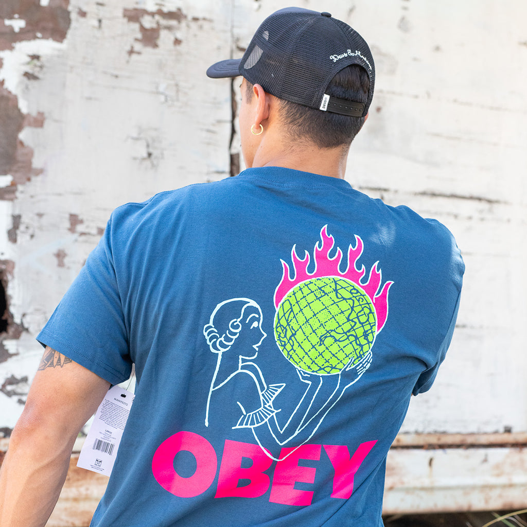 Person wearing a blue t-shirt with a colorful design and 'OBEY' branding, standing against a textured wall.