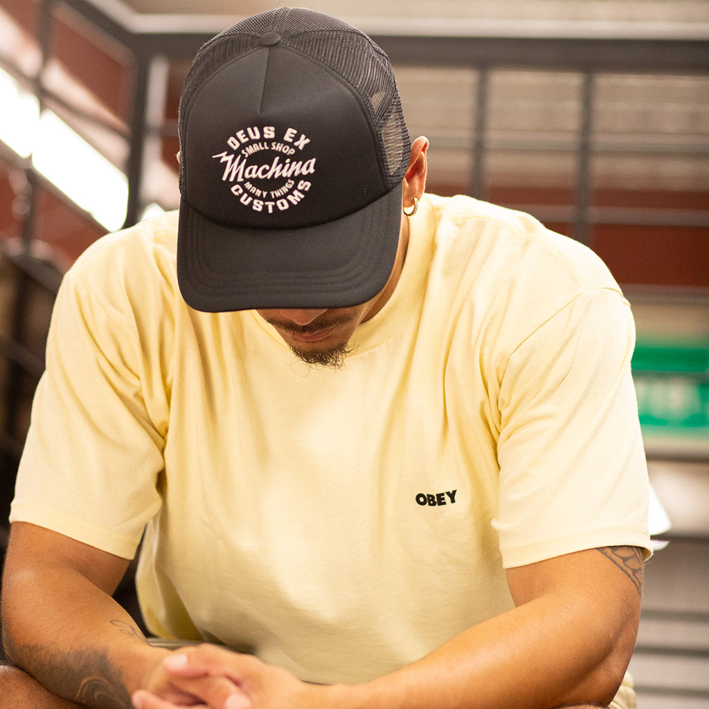 Person wearing a black cap with white text and a yellow t-shirt with 'Obey' logo.