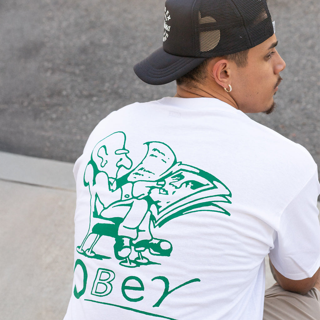 Person wearing a white t-shirt with green graphic design and 'Obey' text, standing against a gray wall.