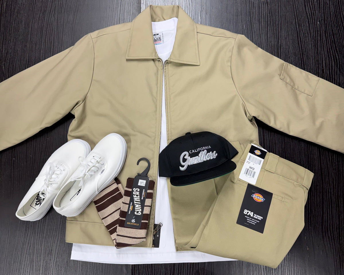 A flat-lay outfit arrangement on a dark wood surface featuring a beige utility jacket, matching beige Dickies pants, white sneakers, striped socks, and a black California Smothers cap. The Dickies brand tag is clearly visible on the pants.