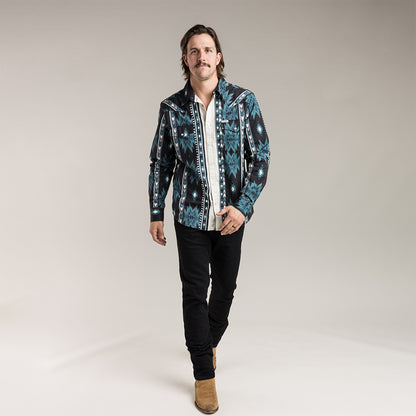 model wearing a patterned button down  over a white shirt with black pants and brown boots on a plain background